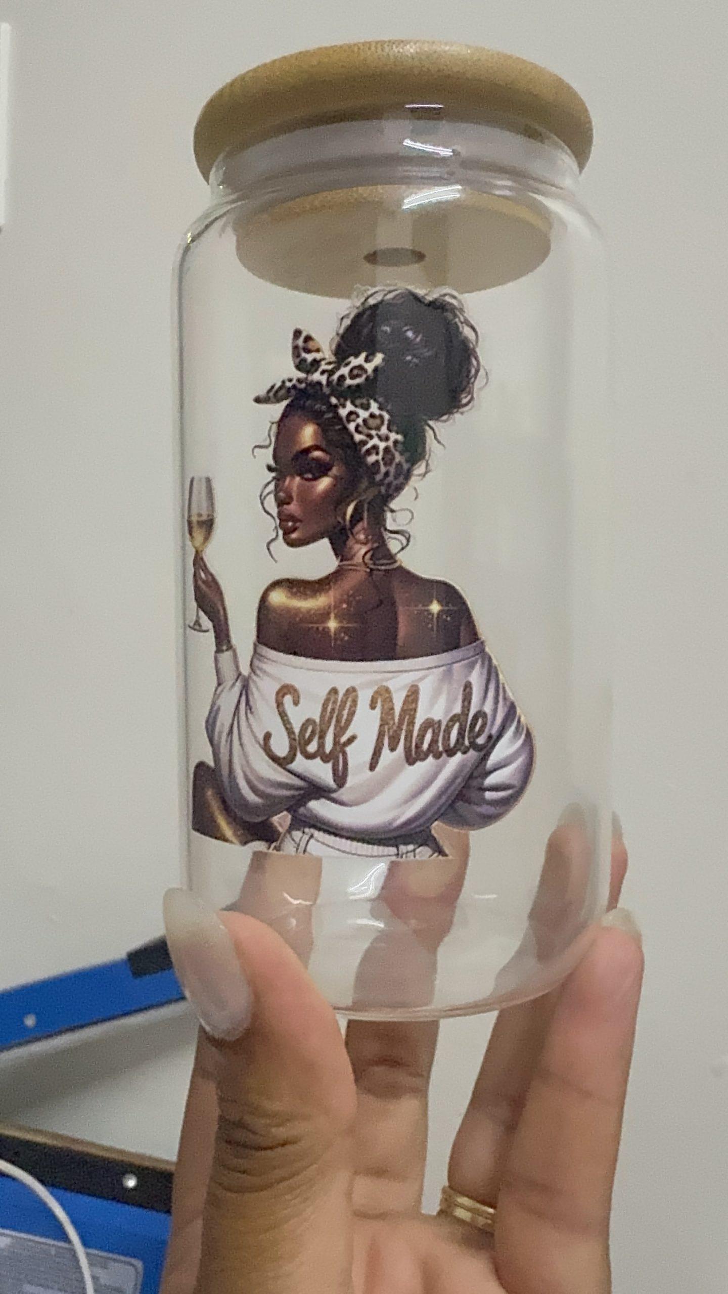 Close-up of a hand holding a clear glass jar with a wooden lid, featuring a detailed graphic of a "Self Made" woman with a champagne glass.